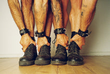 Load image into Gallery viewer, Four lower legs and hands wearing black leather ankle cuffs, each adjusting a black lace-up boot on a light wood floor.